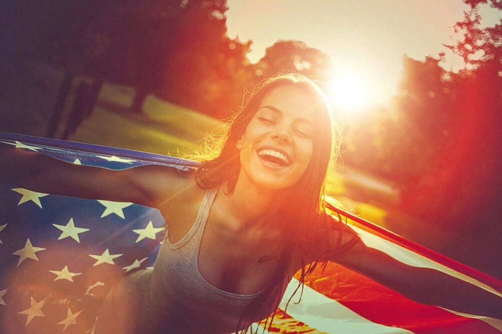 Junge Frau mit US-Flagge im Sonnenuntergang lacht glücklich, Symbol für Freiheit und Gewinnfreude, weil sie Powerball im Lottoland gewonnen hat