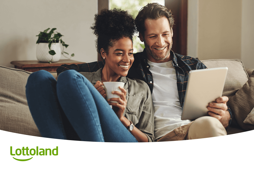 Lächelndes Paar auf dem Sofa entdeckt per Tablet den Lottoland Lotto Quicktipp für den nächsten Jackpot-Gewinn