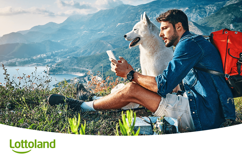 Mann mit Hund sitzt in den Bergen, checkt auf dem Smartphone seine Lottoland Rubbellose – Sommer, Freiheit und Spielspaß vereint