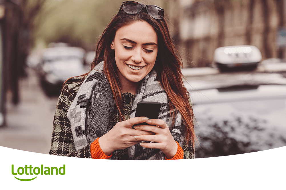 Junge Frau lächelt beim Tippen auf dem Smartphone, unterwegs in der Stadt – Lottospielen mit Lottoland mobil und spontan erleben