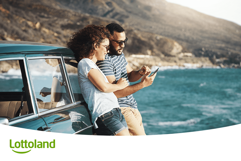 Junges Paar lehnt an Auto und schaut aufs Smartphone – sie prüfen entspannt unterwegs die sichere Millionen-Auszahlung bei Lottoland