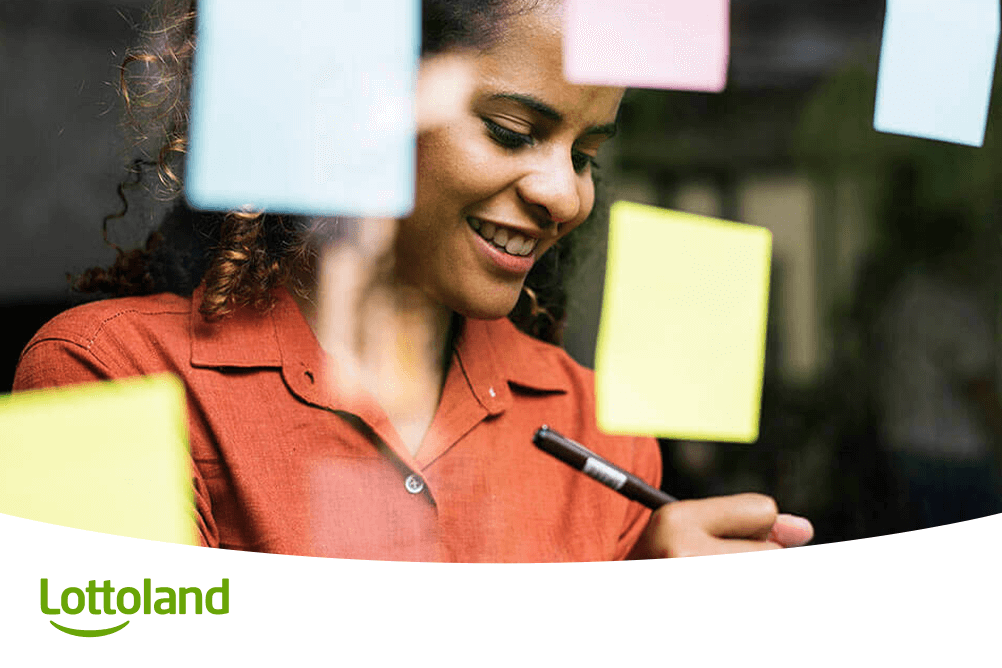 Junge Frau schreibt lächelnd Notizen auf bunte Zettel – sie informiert sich bei Lottoland über die nächste Lottoziehung und plant ihre Tipps